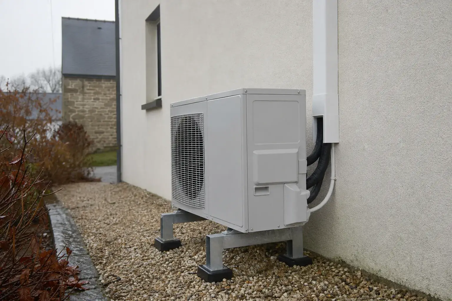 Unité extérieure de pompe à chaleur installée proprement contre la façade enduit clair d'une maison bretonne, tuyauterie gainée